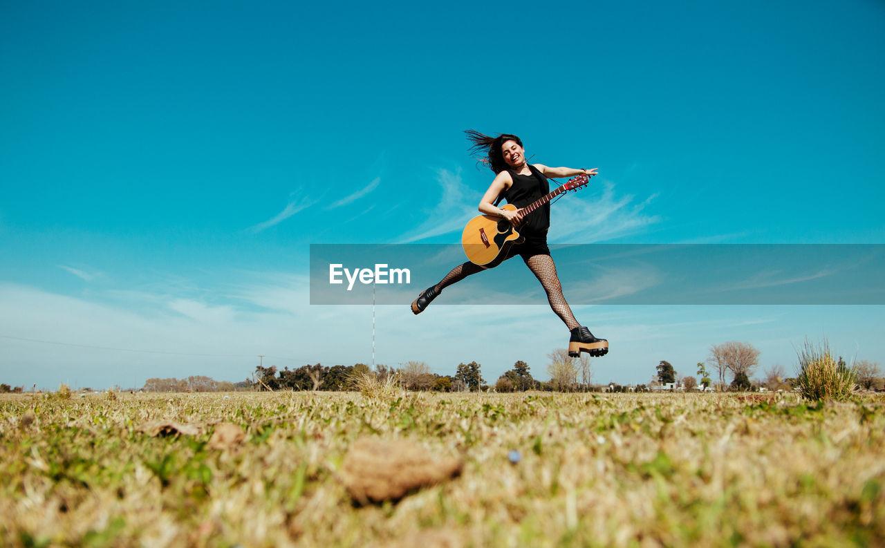 Woman with guitar jumping over field against sky