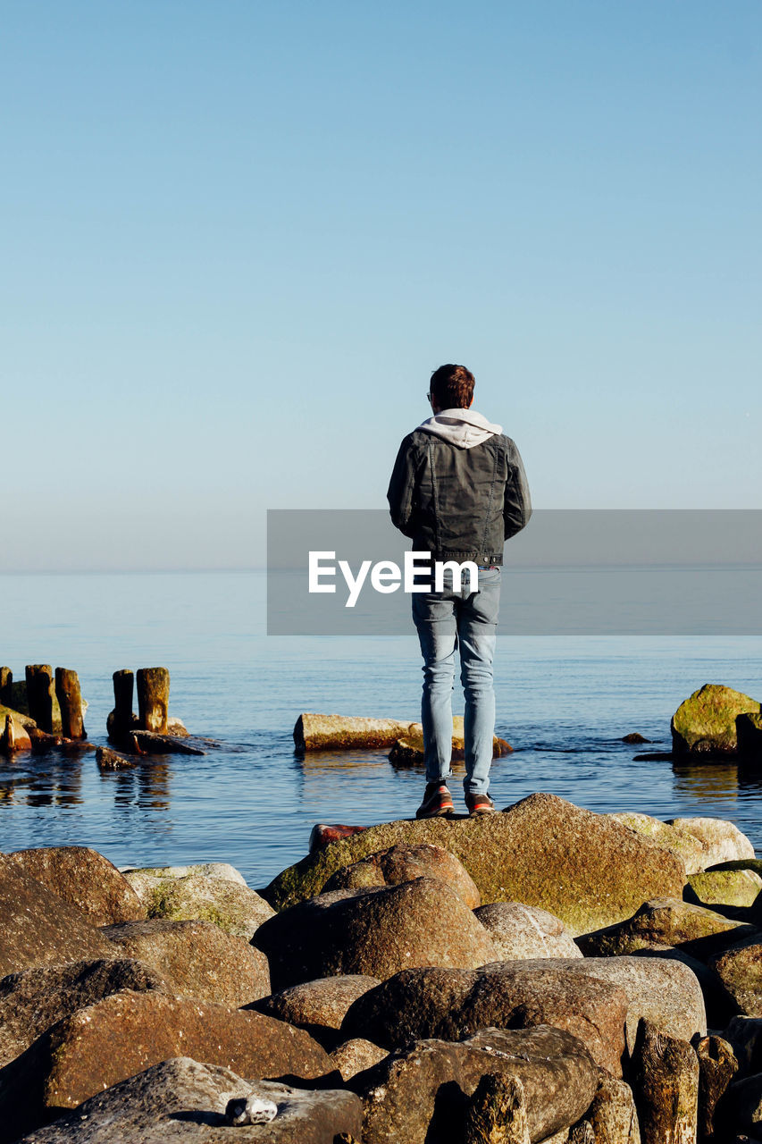 Full length rear view of man standing on rocks by sea against clear sky