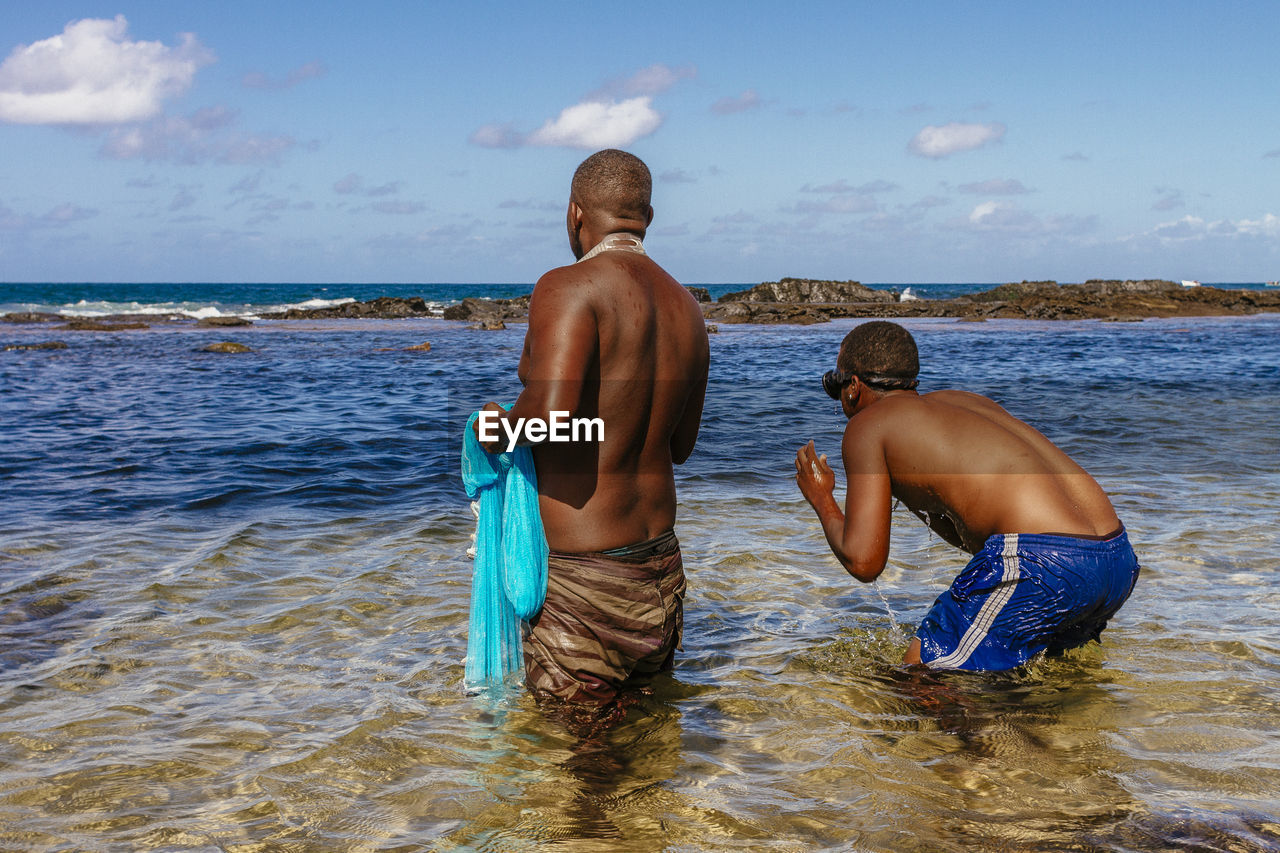 Shirtless fishermen standing in sea against sky