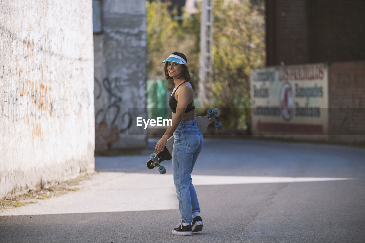 Side view full length portrait of young woman standing with skateboard on street