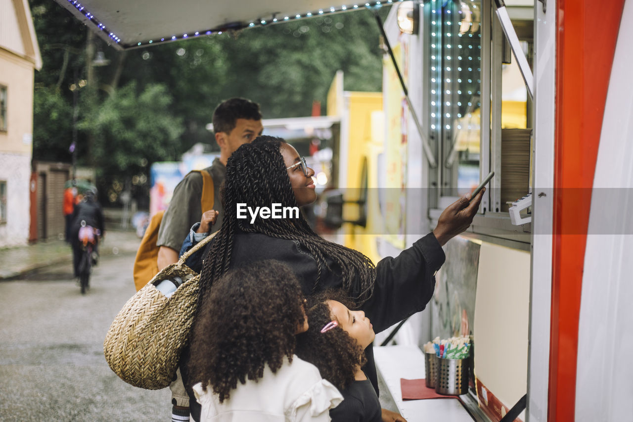 Smiling woman doing online payment near food truck while standing with family at amusement park