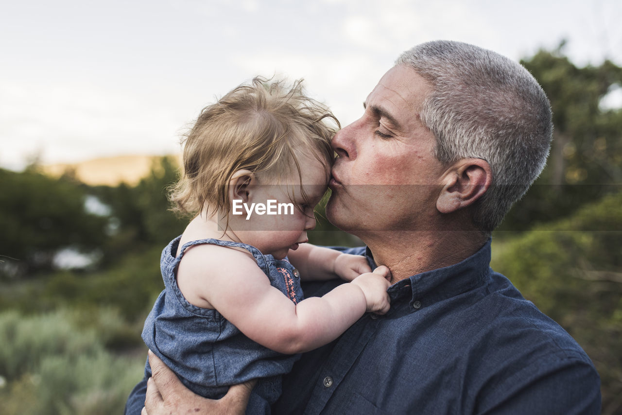 Close-up of father kissing on daughter's forehead while standing against sky in forest