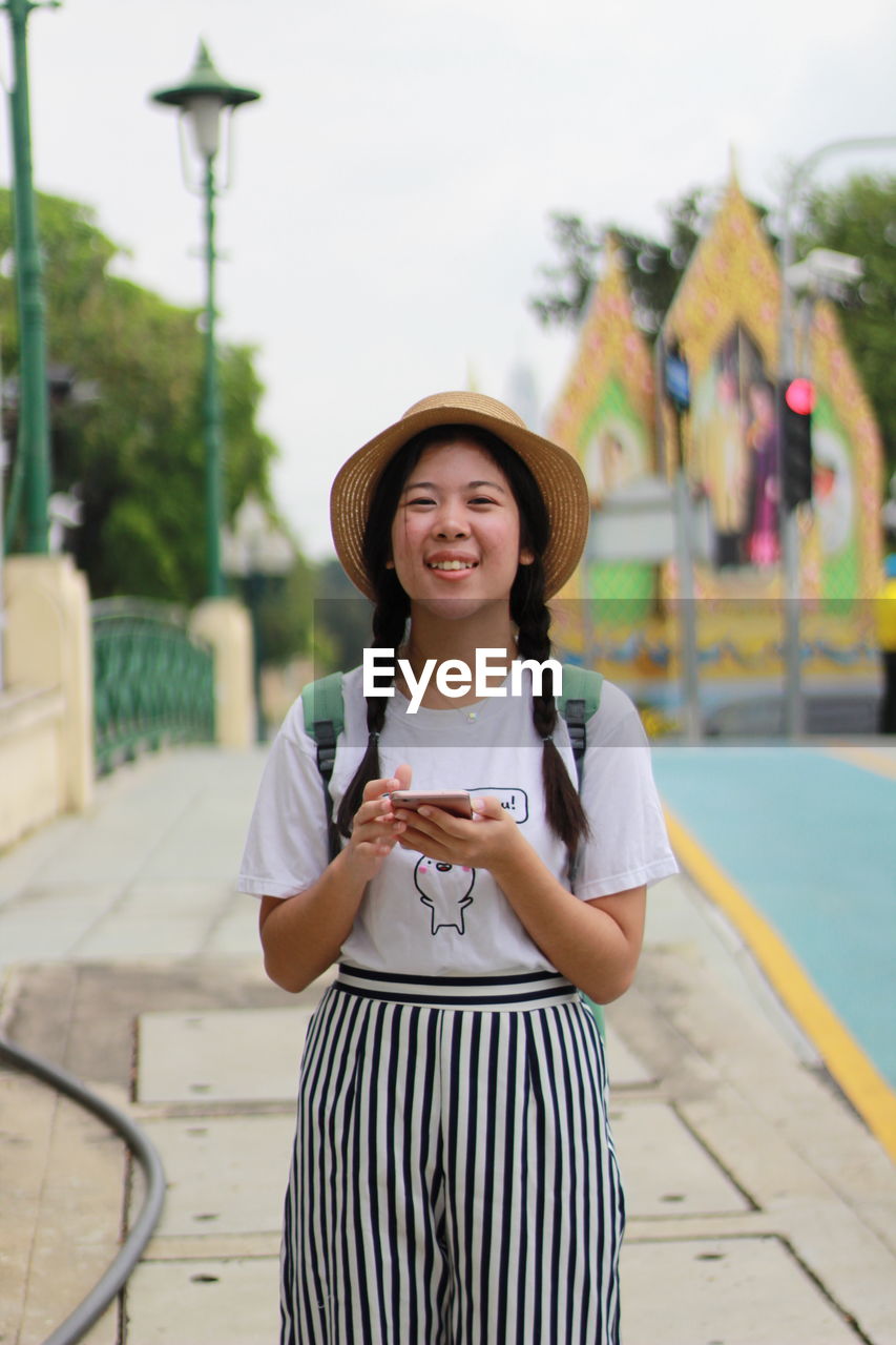 Portrait of smiling woman using mobile phone while standing against sky