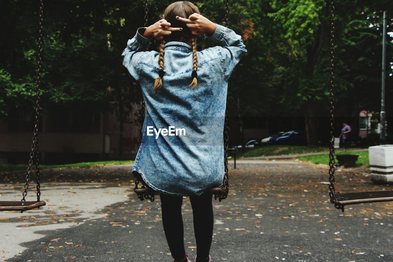 Rear view of woman showing middle fingers while sitting on swing at park