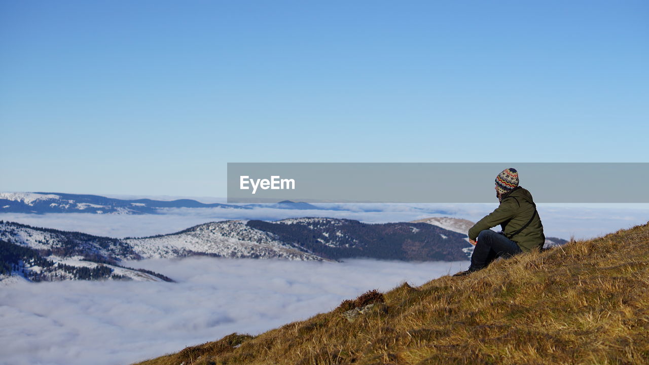 Man looking at view while sitting on mountain against clear sky