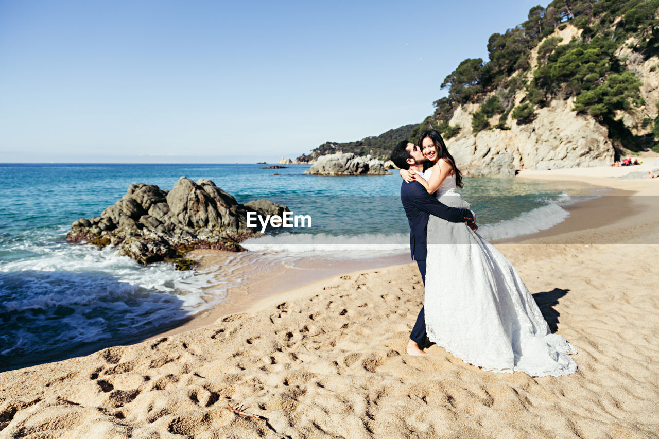 Happy newlywed couple romancing at beach against clear sky