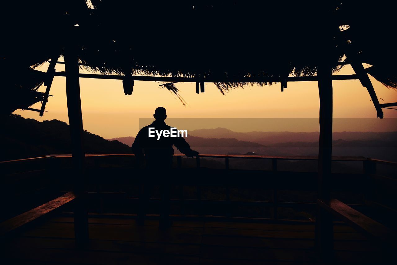 Rear view of silhouette man standing by railing against sky during sunset