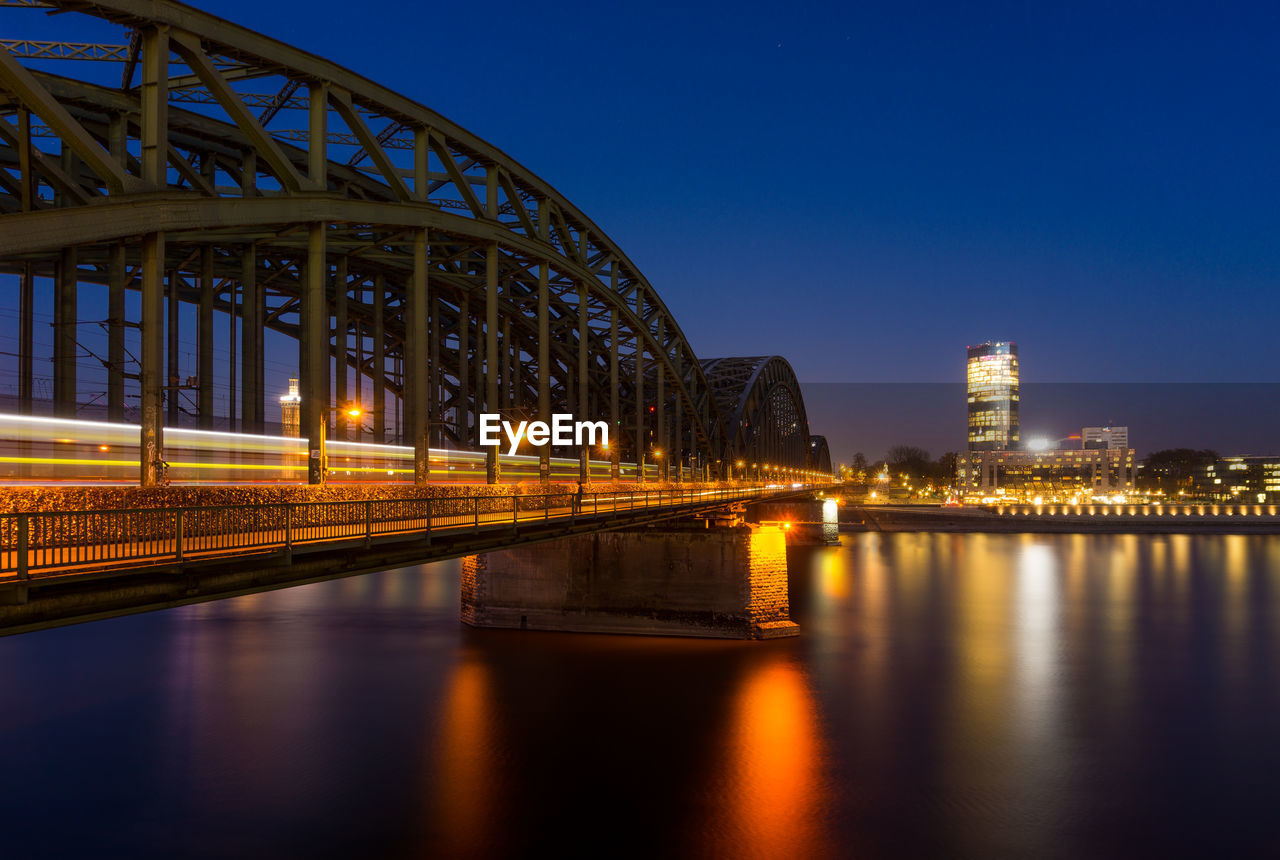 Illuminated bridge over river by buildings against sky at night