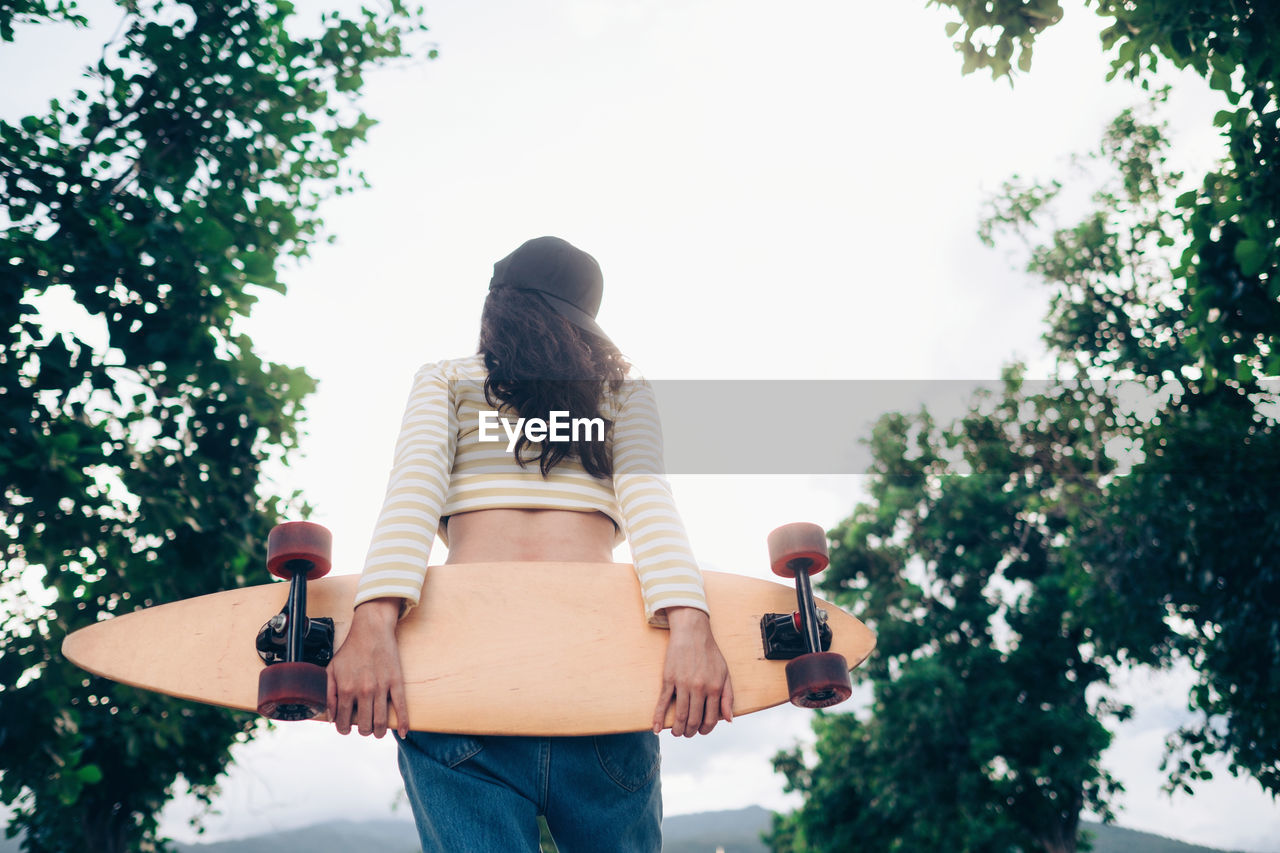 Rear view of woman holding skateboard while standing against trees