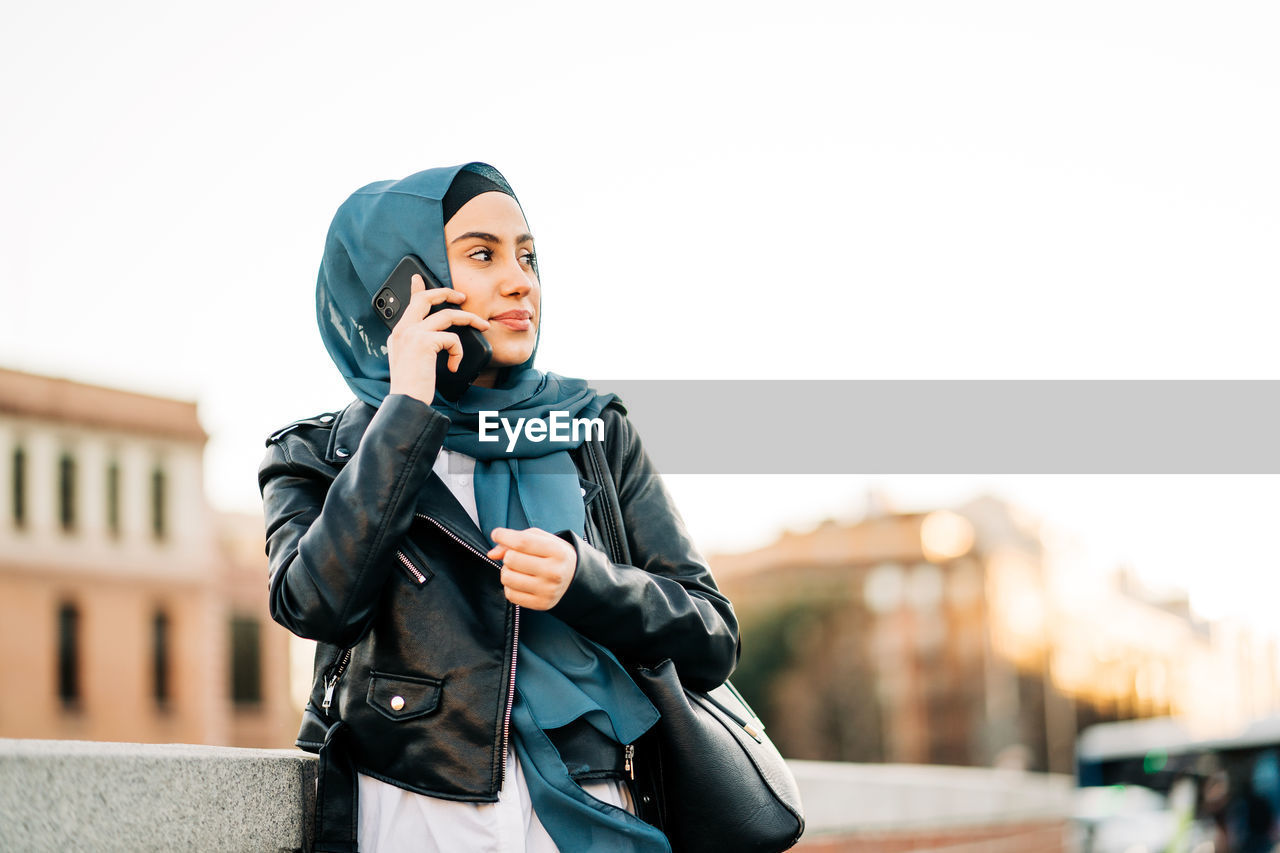 Cheerful muslim female in traditional headscarf standing on city street and speaking on mobile phone while looking away