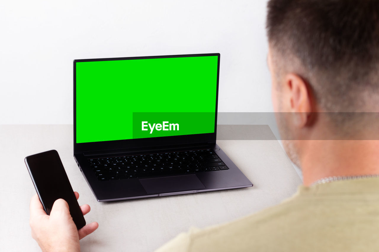 A man with a phone in his hand is sitting in front of a laptop with a green mockup on the monitor