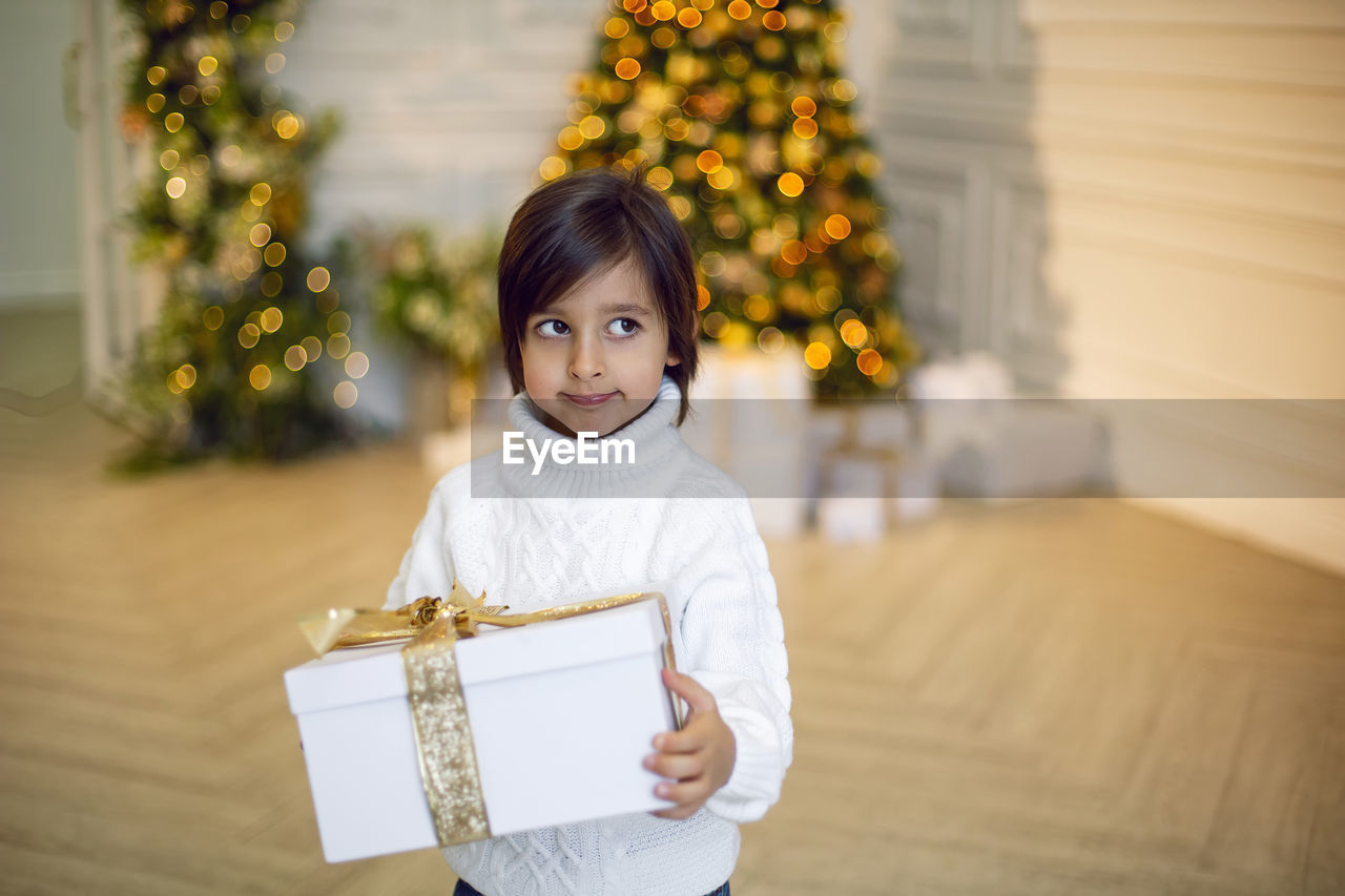 Portrait of a boy a child in a white sweater holding a gift box at the christmas tree