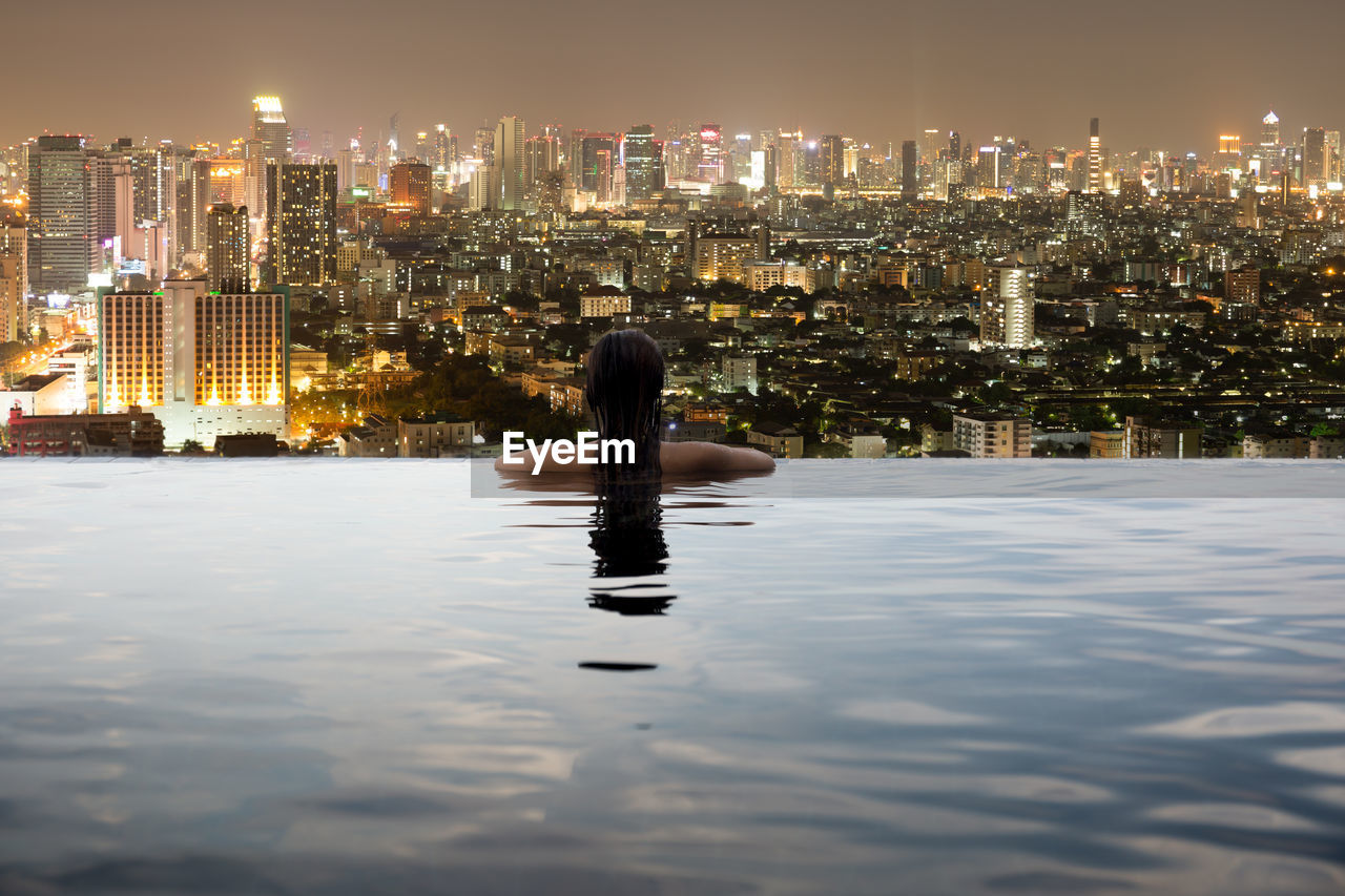 Rear view of woman in swimming pool against illuminated city at night