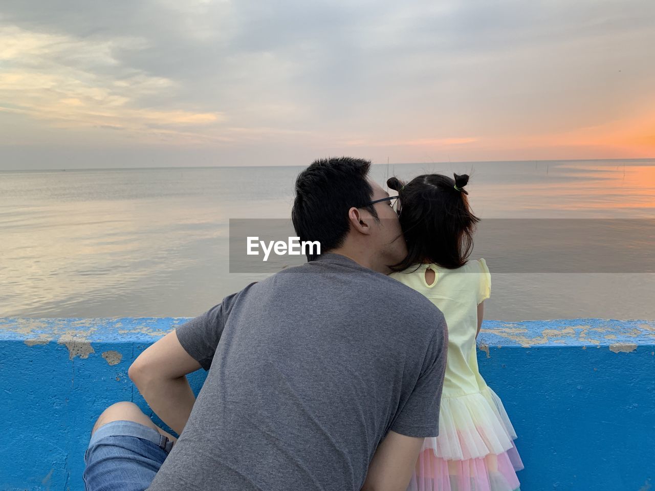 Rear view of father kissing daughter against sea during sunset