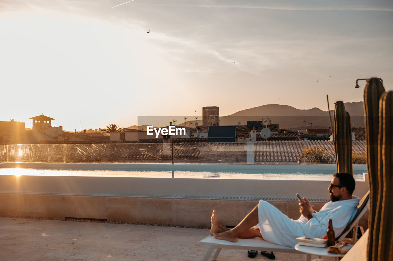 Side view of young bearded male traveler in white robe and sunglasses relaxing on lounger on rooftop and messaging on smartphone against sunset sky during vacation in mallorca