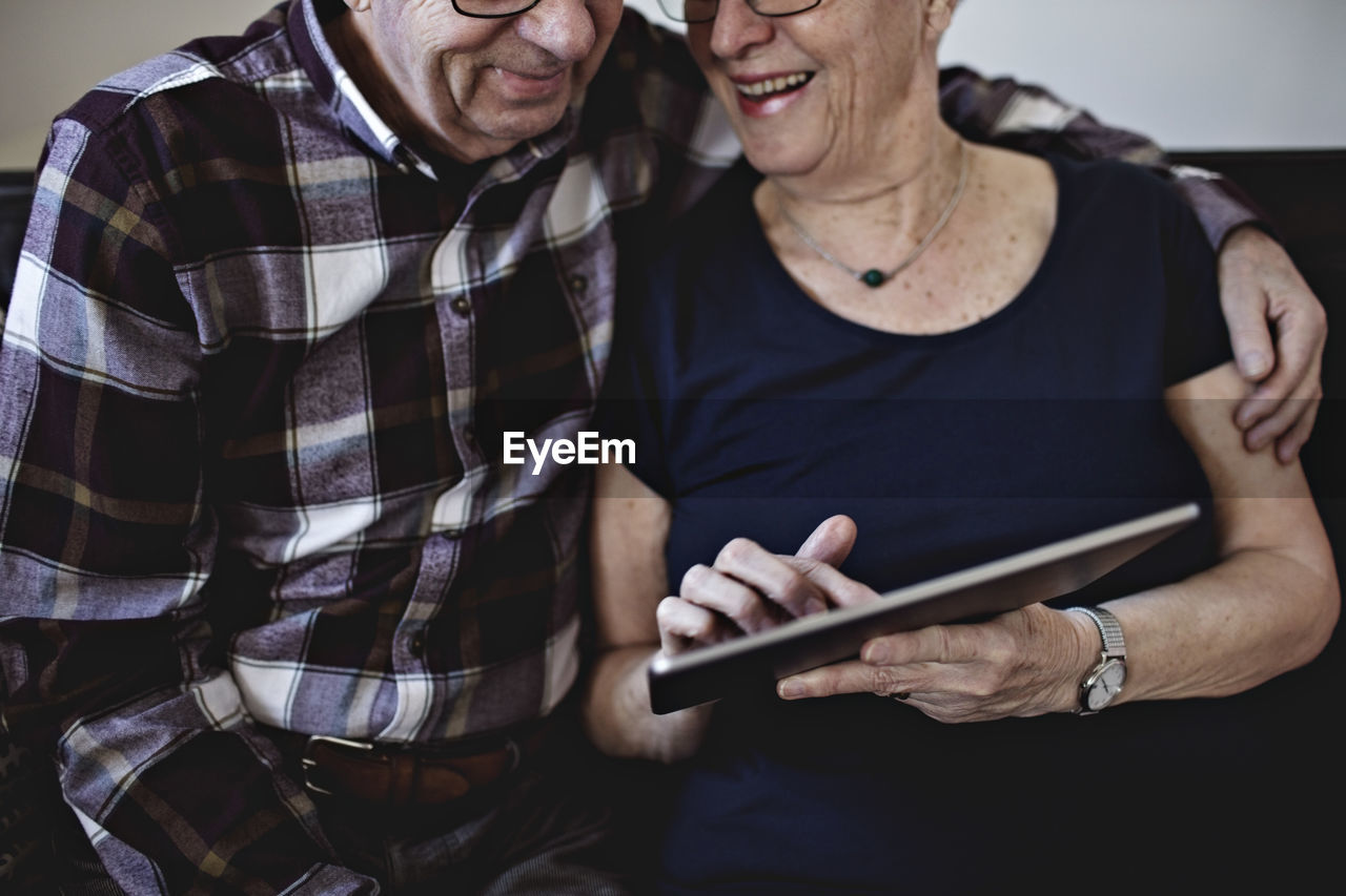 Midsection of smiling senior couple sitting while sharing digital tablet at home