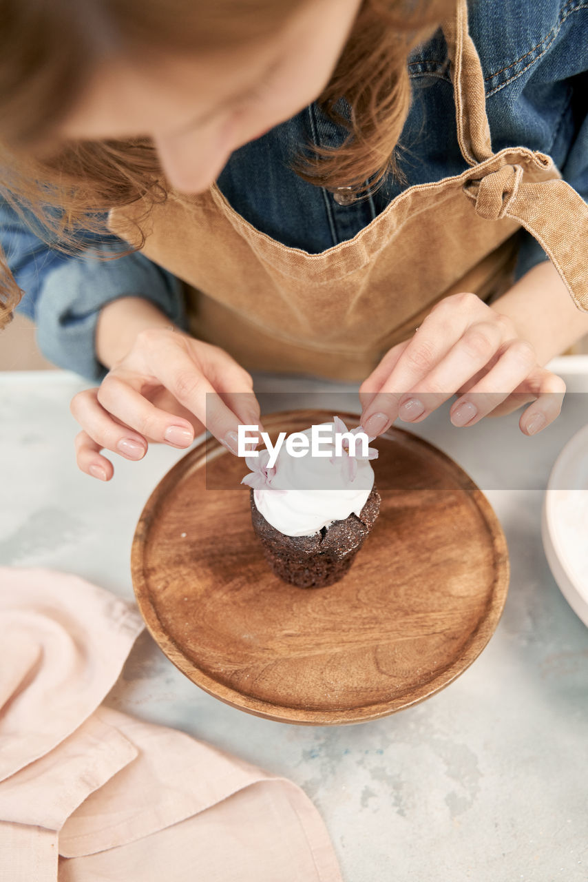 Crop young female decorating chocolate cupcake with sweet whipped cream and flowers on cake stand while cooking at home