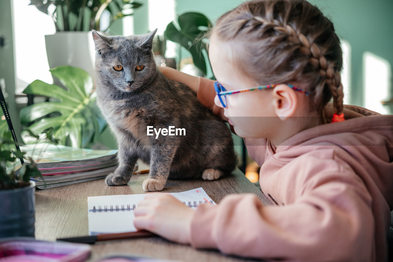 Little girl in eyeglasses with her pet cat having online lesson during covid lockdown, distance