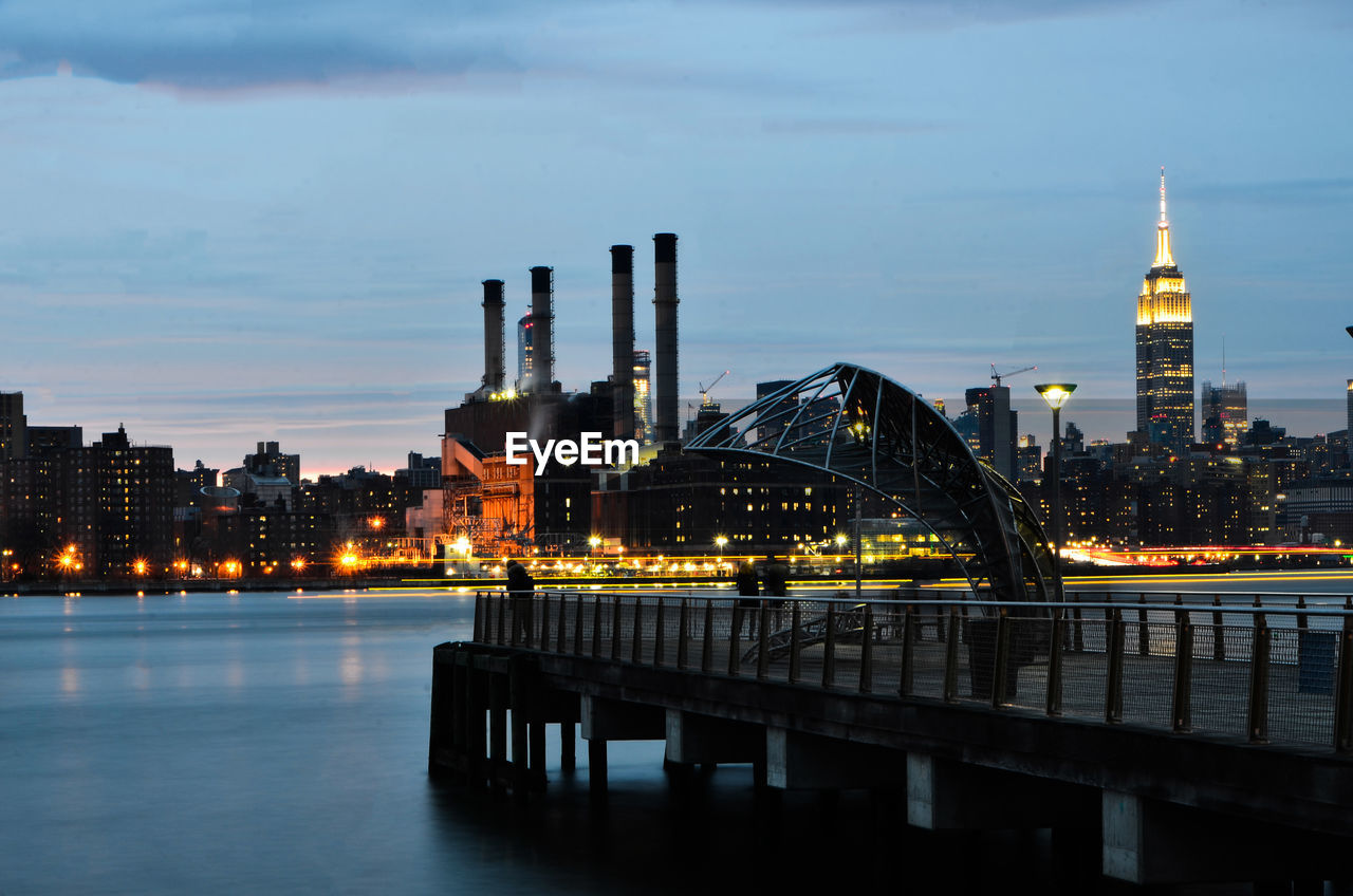 Illuminated bridge over river by buildings against sky at night.