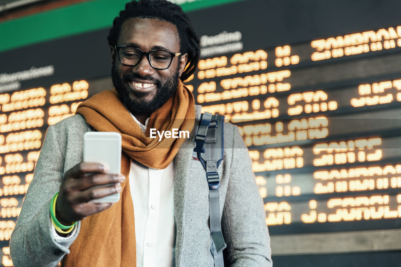 Smiling man using mobile phone while standing airport departure board