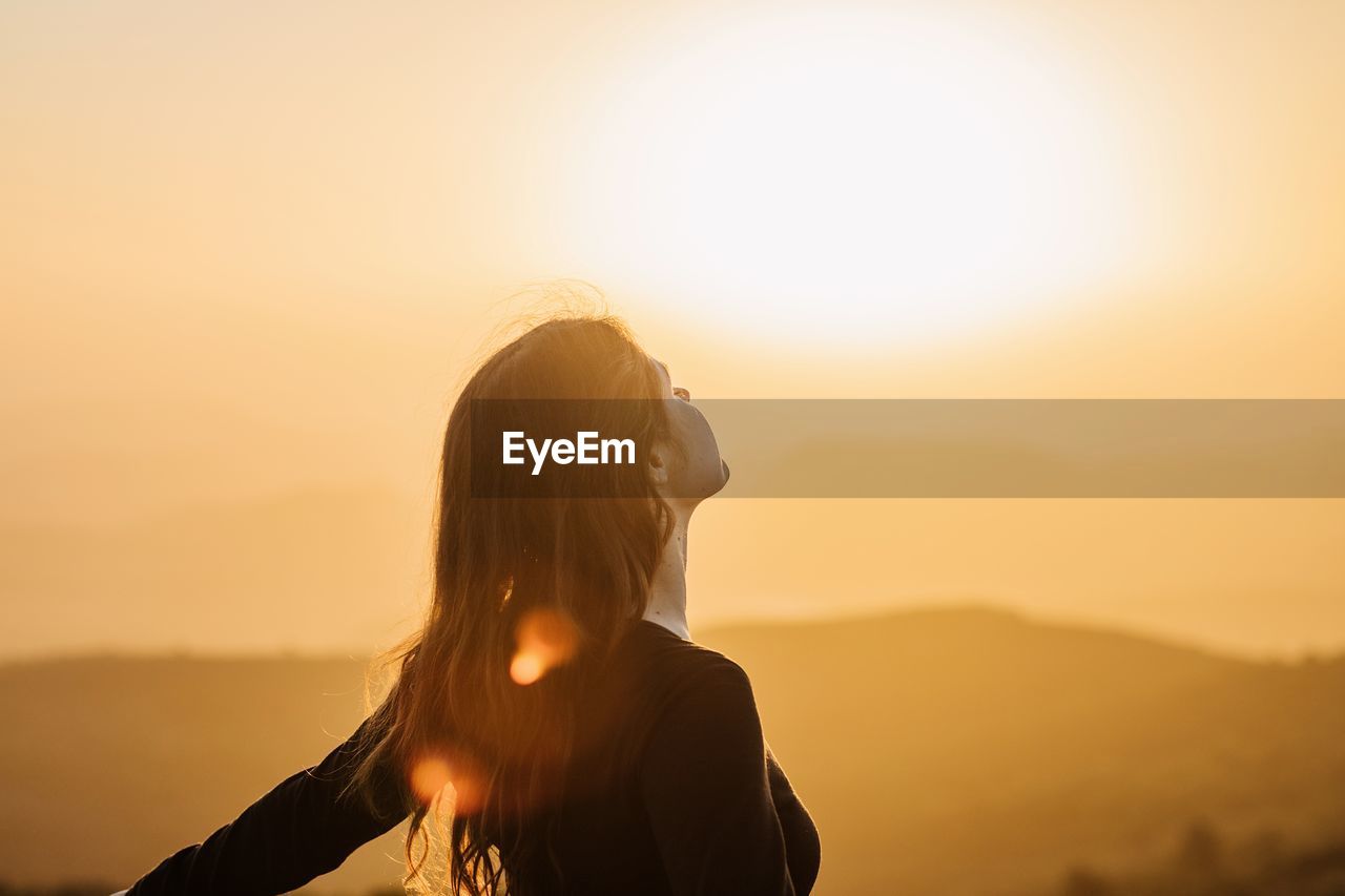 Side view of happy female standing with eyes closed on hill and enjoying freedom while admiring mountainous scenery at sunset