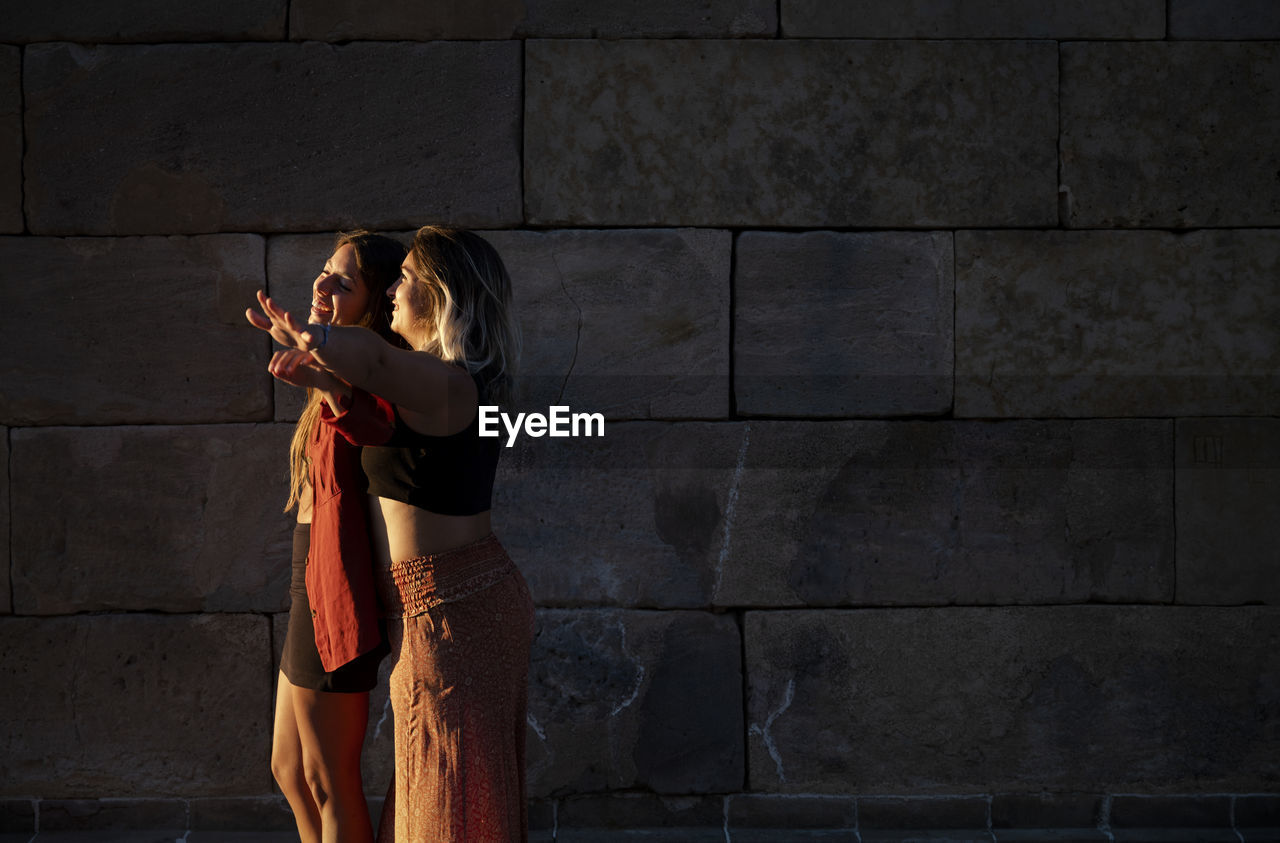 Two girls standing against stone wall during sunset