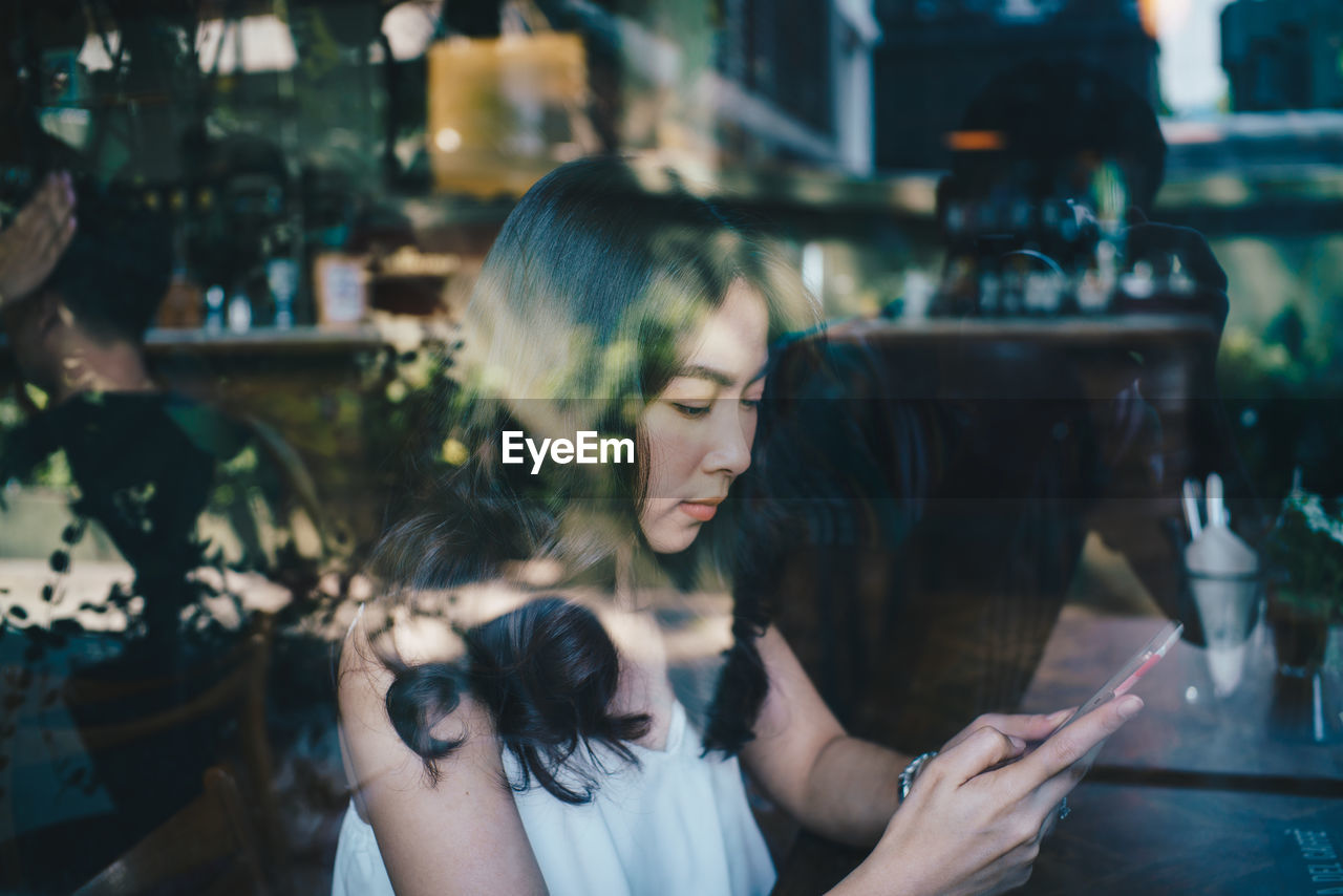 Side view of woman using mobile phone while sitting at table seen through glass window