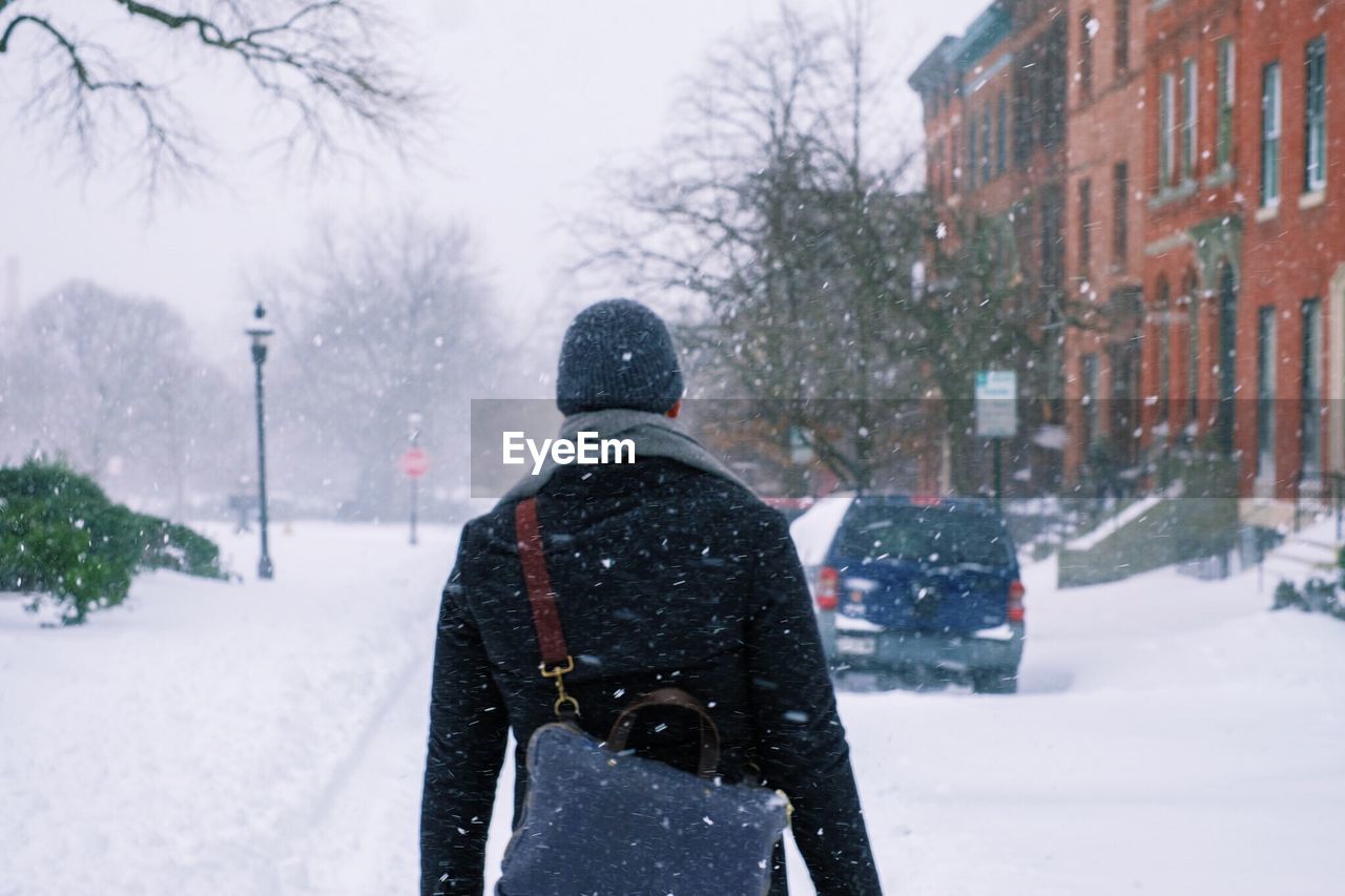 Rear view of man with bag walking on snow covered field in city