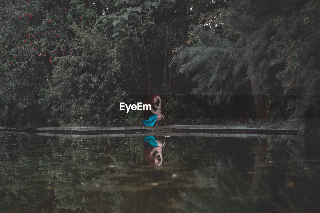 Side view of teenage girl crouching by lake at forest