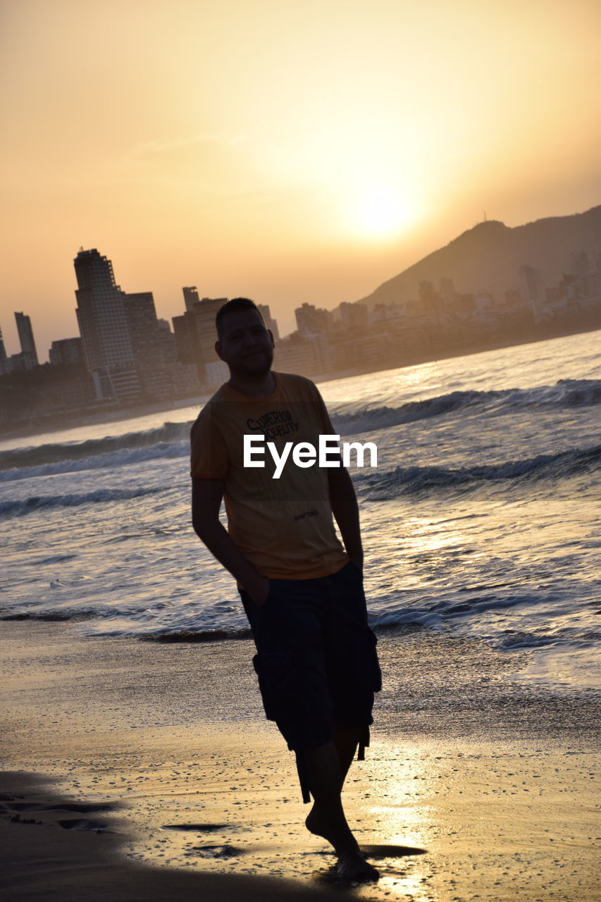 FULL LENGTH OF MAN STANDING AT BEACH DURING SUNSET