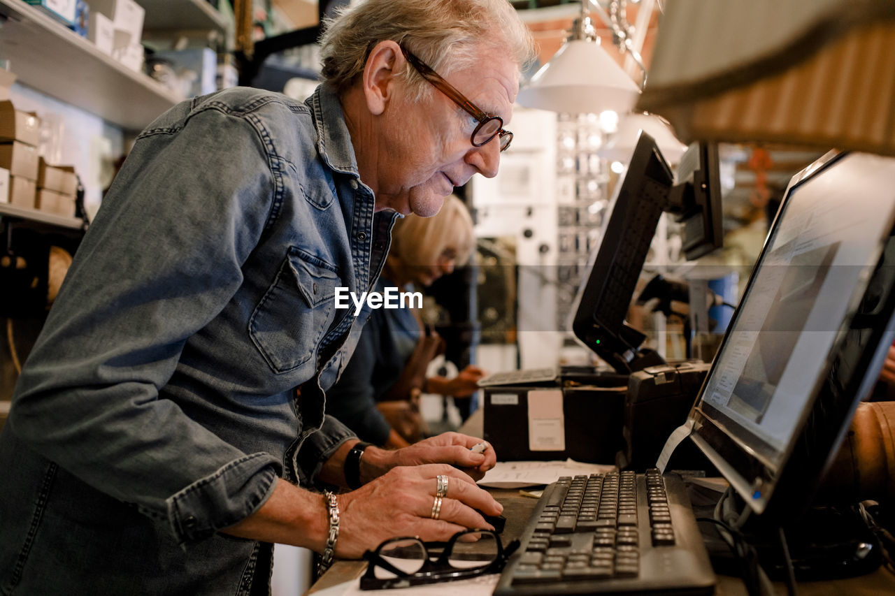 Side view of confident salesman in denim shirt using computer at hardware store checkout