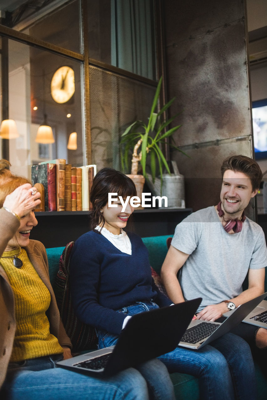 Smiling male and female colleagues looking at female manager sitting with laptop on sofa at creative office