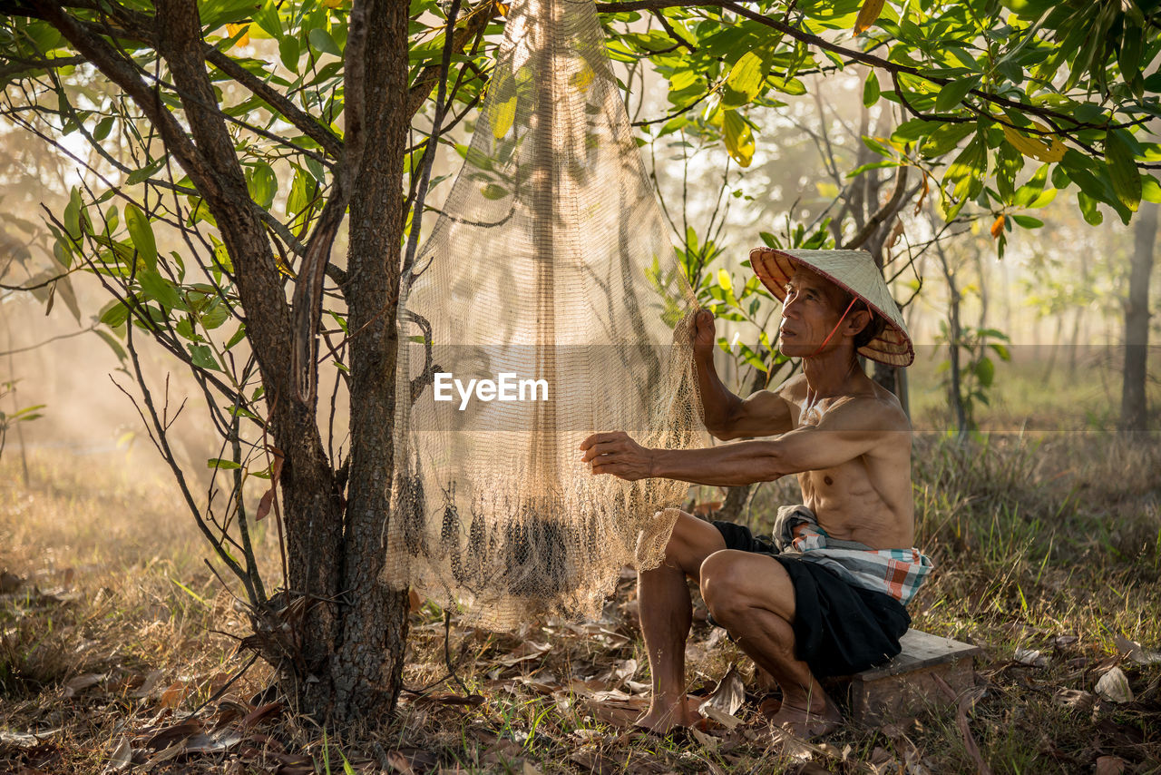 Shirtless senior man drying fishing net at forest