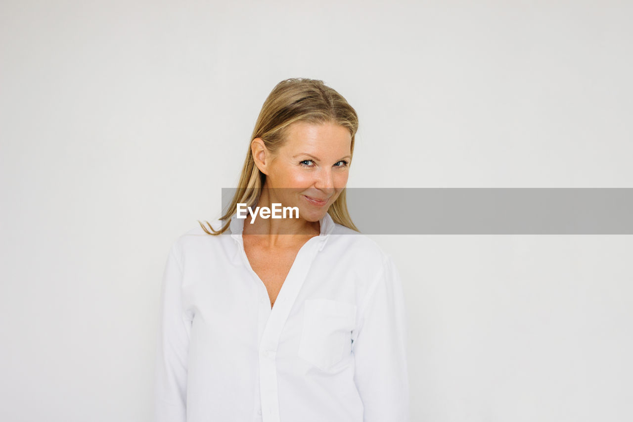 Portrait of flirting blonde forty year woman with long hair in white shirt on the white wall 