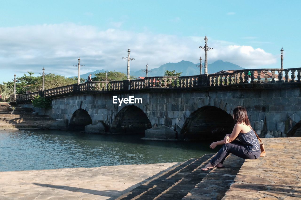 Side view of woman sitting on promenade besides bridge by river