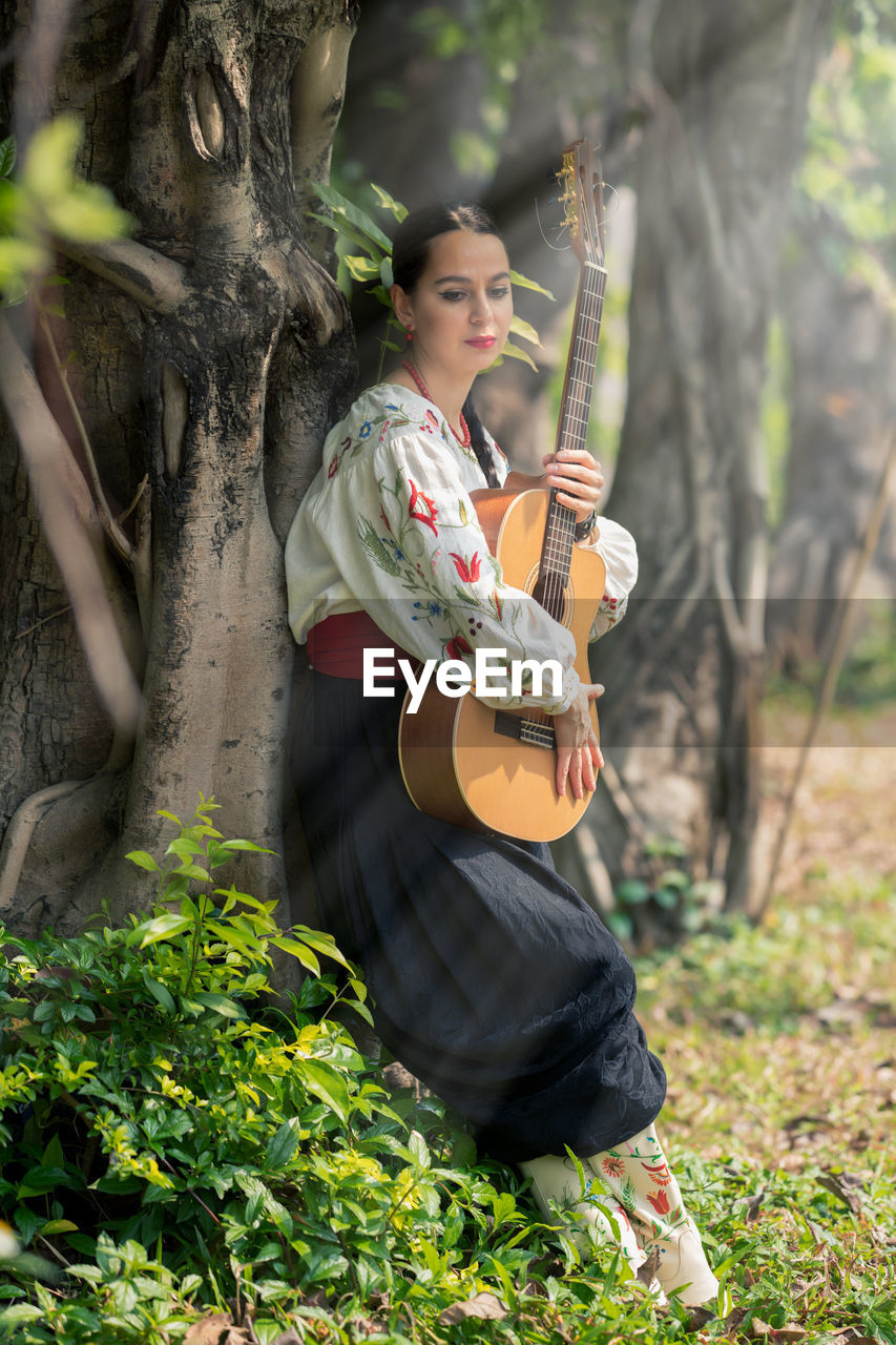 Young woman with guitar leaning on tree at forest