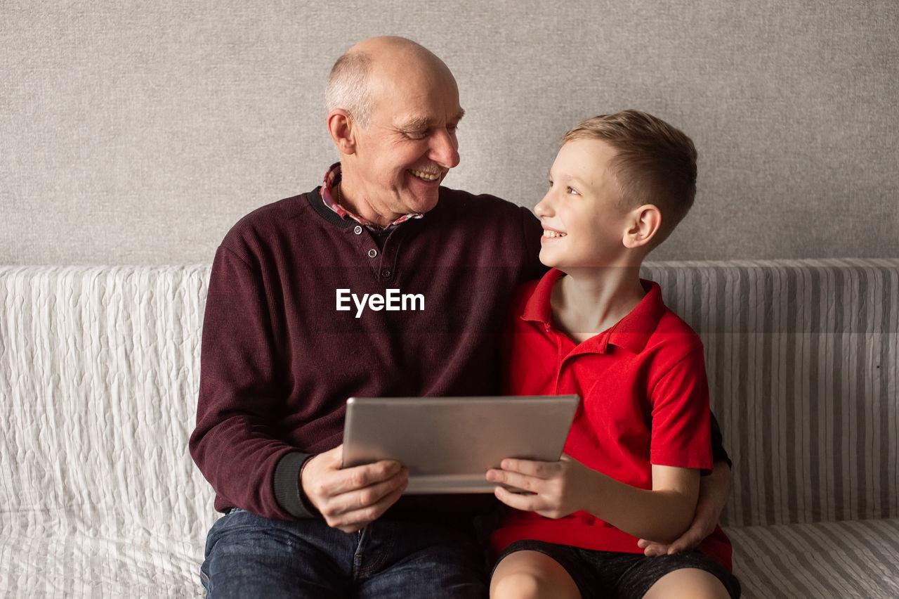 A happy grandfather hugs his grandson. they hold a tablet in their hands and look at each other