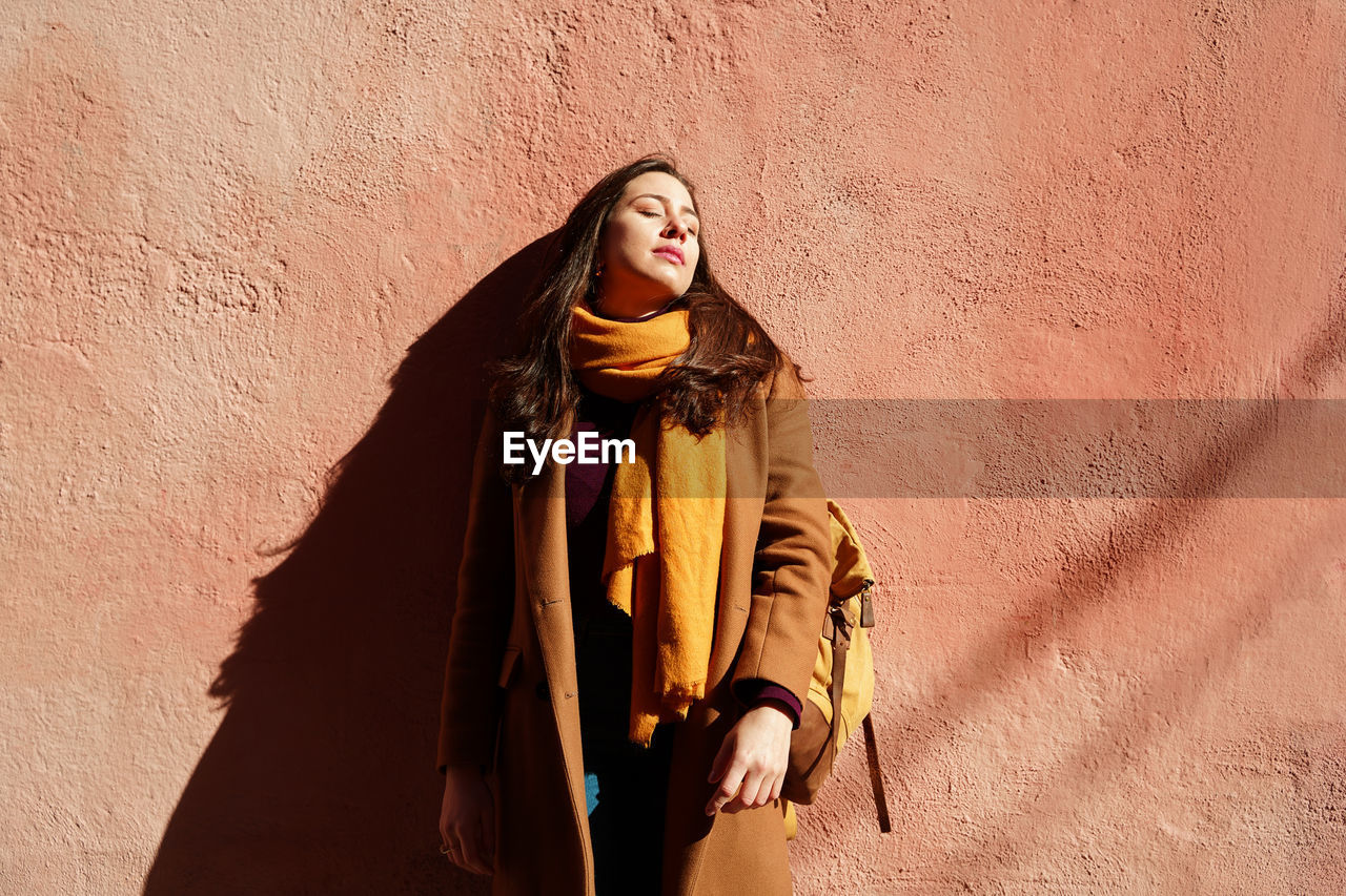 Young concentrated female in outerwear with rucksack and closed eyes standing on walkway in sunlight on coral background