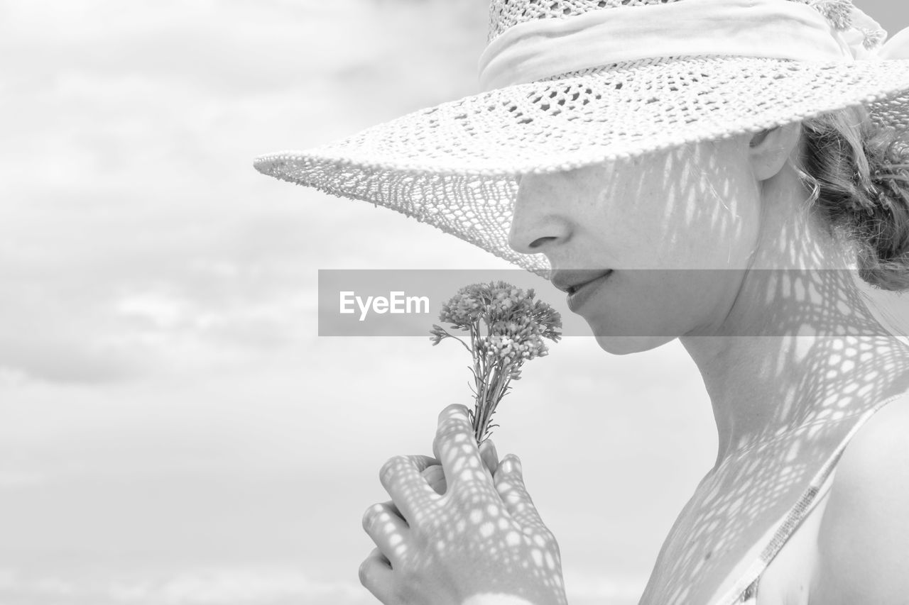 Close-up of woman smelling flower wearing hat against sky