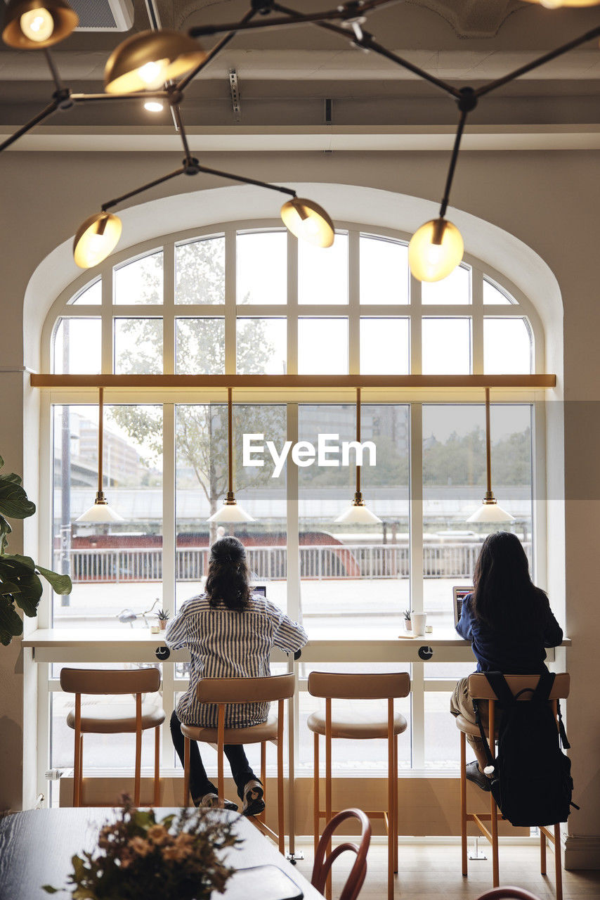 Rear view of businesswomen working on laptop while sitting on chair near window at coworking office