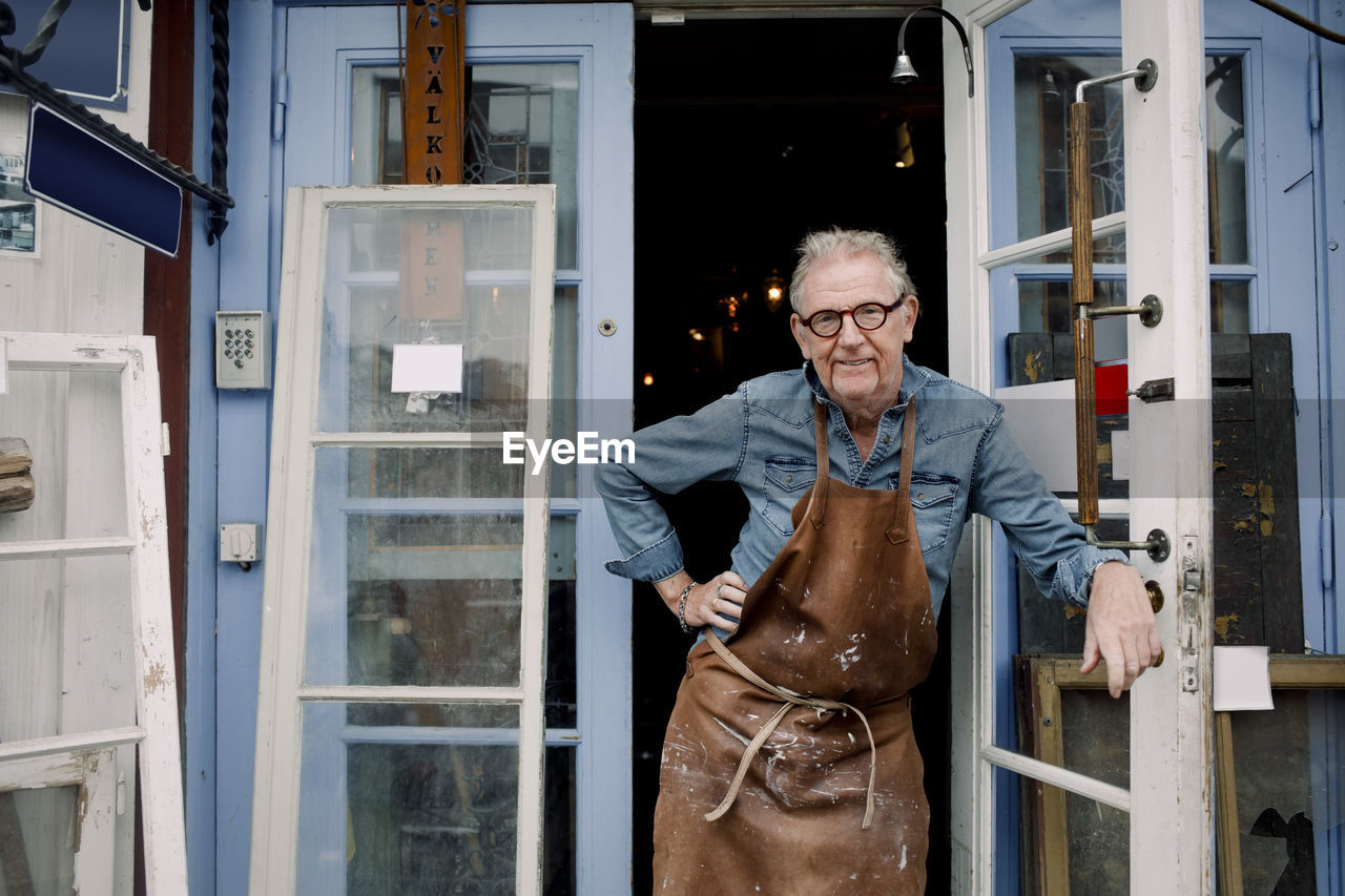 Portrait of senior hardware store owner standing with hand on hip while leaning on door