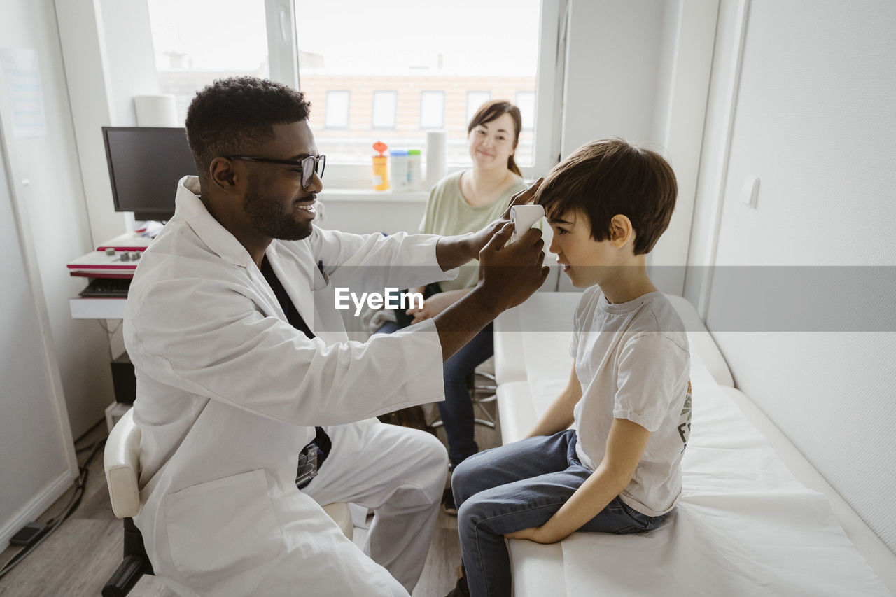 Male pediatrician examining boy sitting on bed in examination room at healthcare center