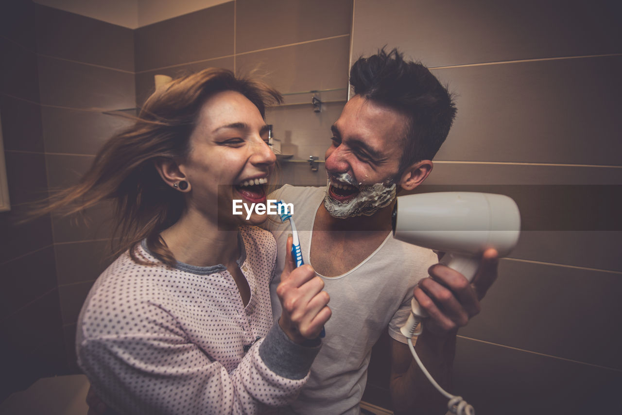 Woman singing while man holding hair dryer in bathroom