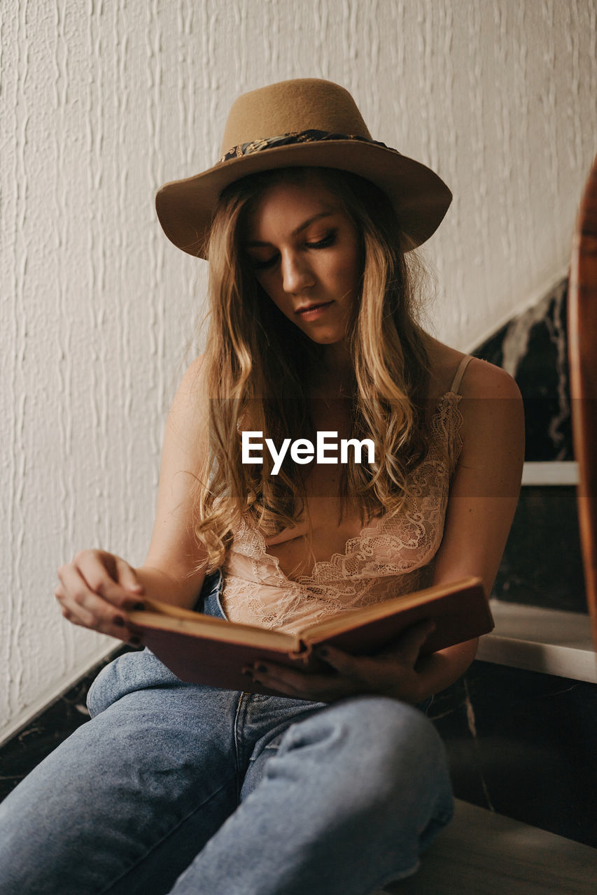 Pensive female wearing hat and jeans sitting on stairs in modern house and reading interesting book while relaxing at weekend at home