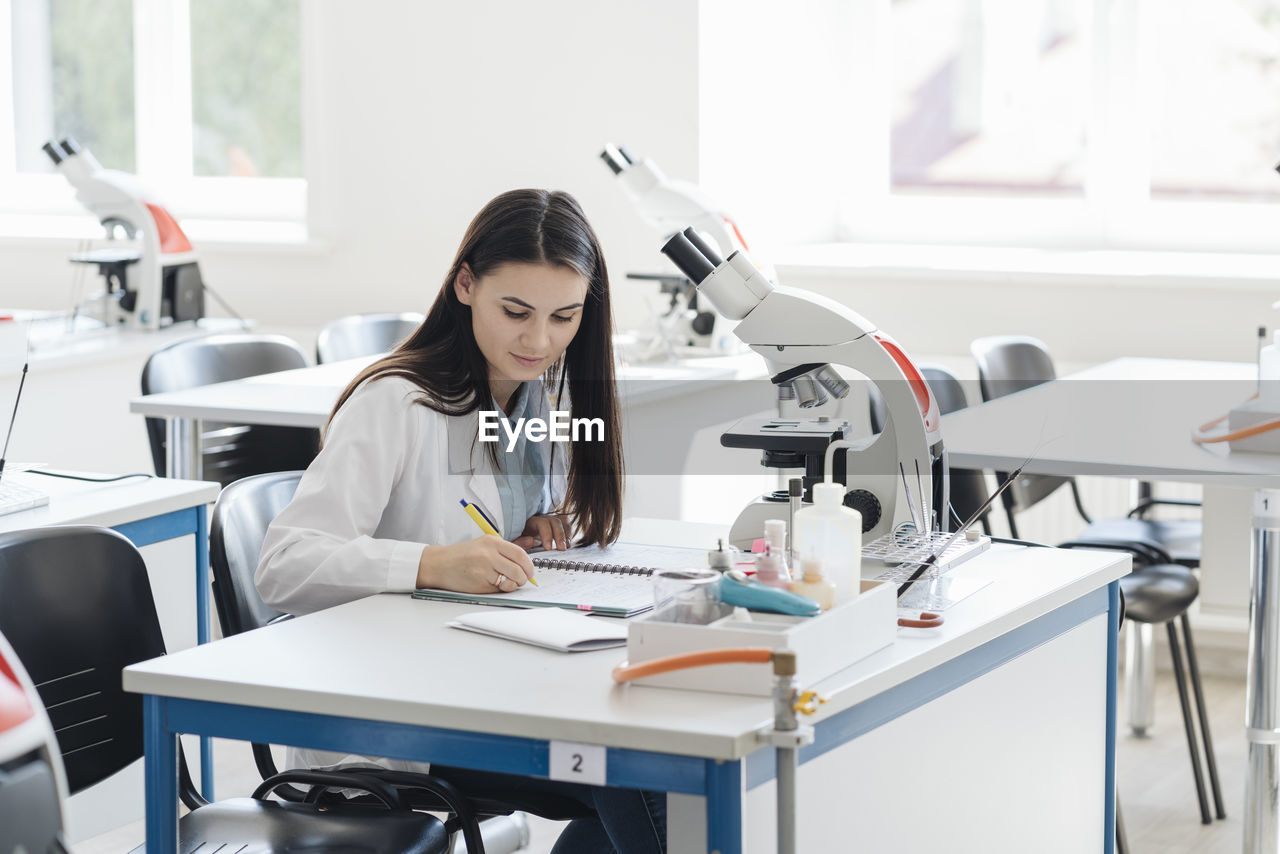 Young researcher in white coat taking notes in science class