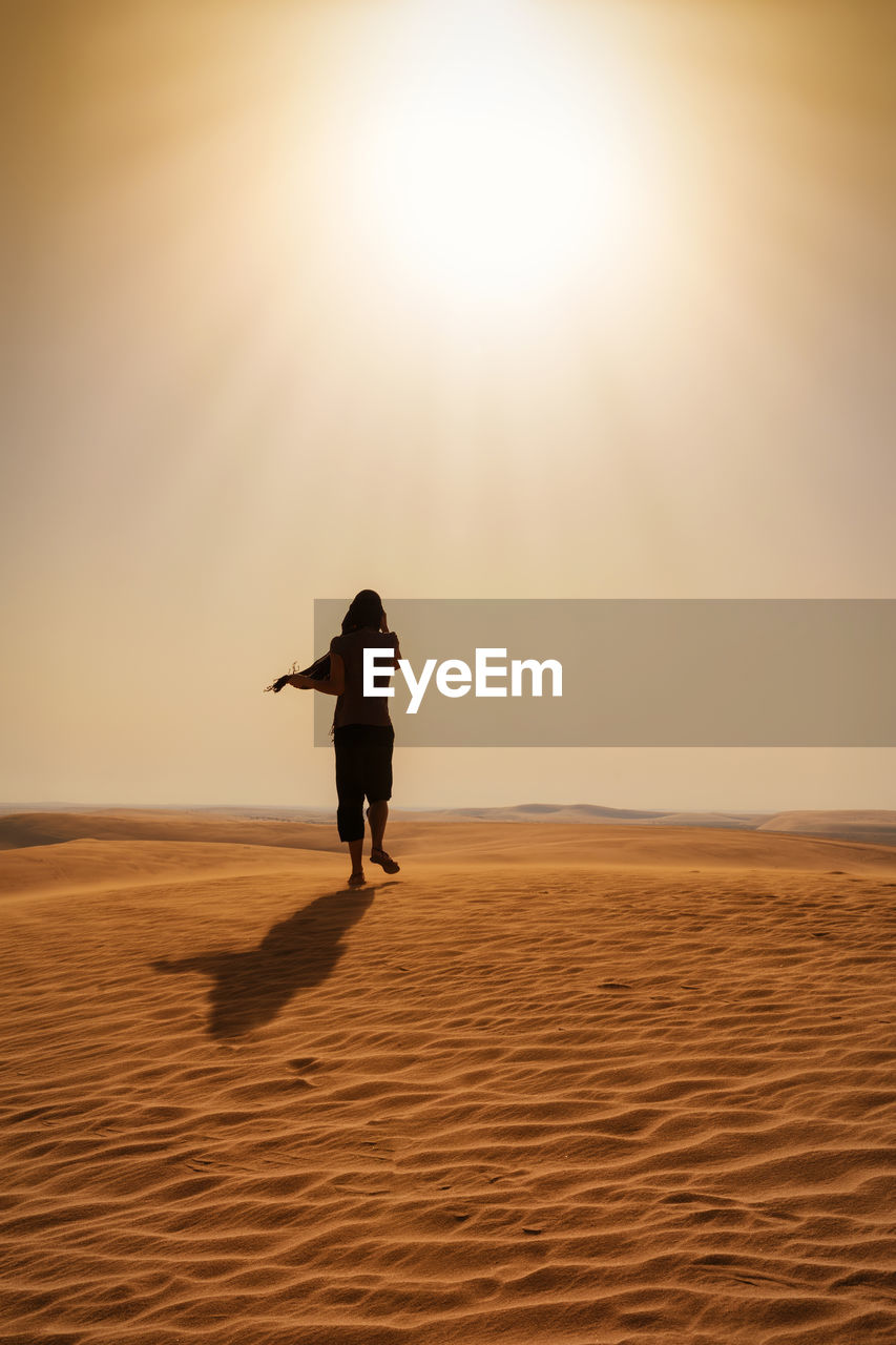 Rear view of woman walking on sand at desert against sky during sunny day