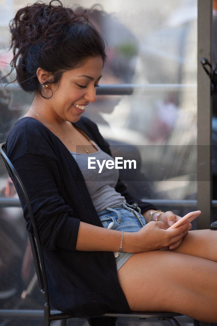 Young woman using mobile phone while sitting by window at restaurant