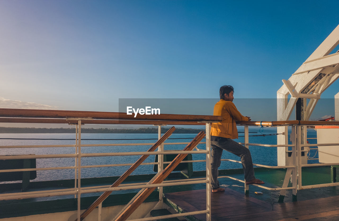one person, sky, men, architecture, water, adult, railing, vacation, nature, full length, standing, blue, sea, clear sky, rear view, copy space, built structure, young adult, casual clothing, sunlight, day, outdoors, lifestyles, travel, occupation, wood, person, leisure activity, transportation, looking, trip, sunny