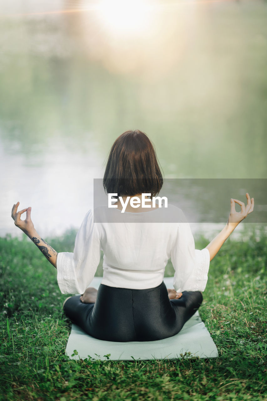 Young woman sitting in lotus position and practicing meditation near water in the nature