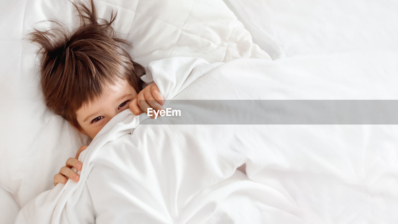 Cute toddler hides in bed. disheveled boy wakes up and peeps out from under white blanket. morning.